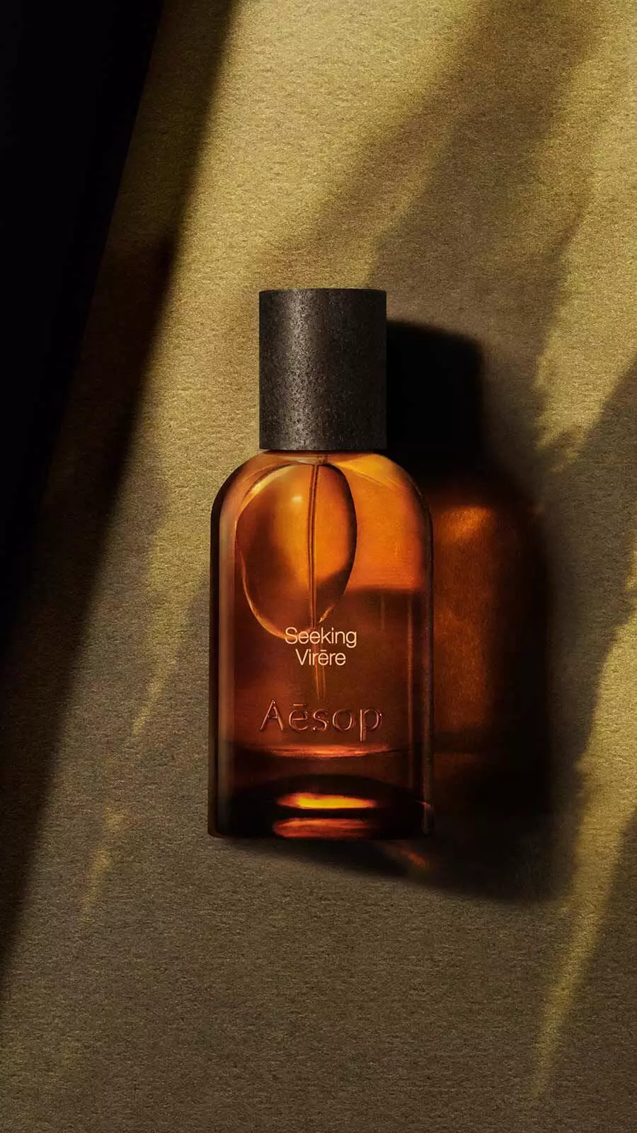 Aesop Seeking, Virere Eau de Parfum bottle with black cap on a warm golden-green surface with dramatic diagonal shadow lines.