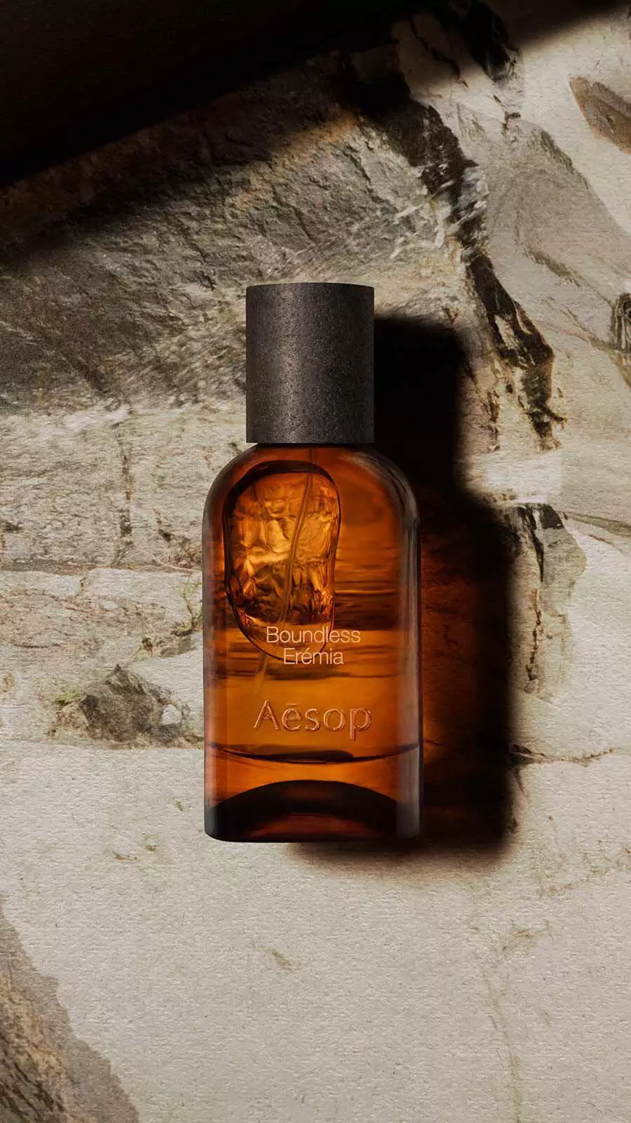 Aesop Boundless, Eremia Eau de Parfum bottle with black cap resting against a weathered pale stone wall in warm directional light.