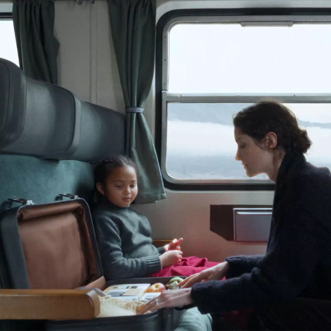 An adult leans over an open suitcase containing fruit and boxes beside a child seated in a green-upholstered train compartment.