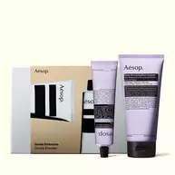 Two lilac Aesop tubes with black caps stand in front of a silver gift box with a gold illustrated sleeve.